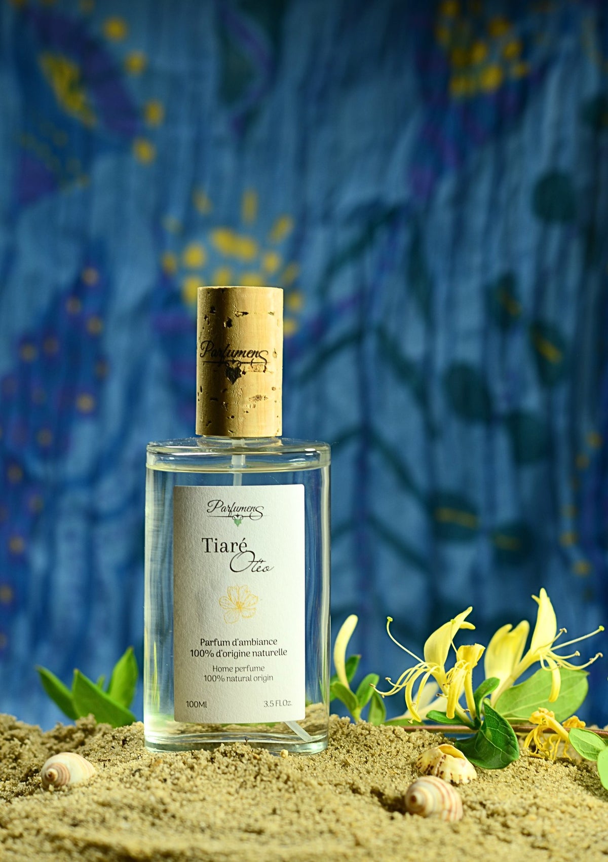 Flacon Tiaré Otéo de Parfumens posé sur le sable, entouré de fleurs
